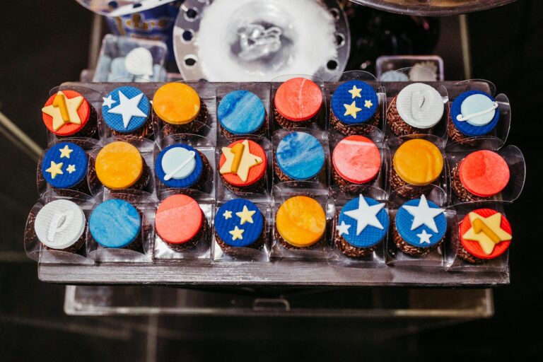 Assorted space-themed cupcakes with colorful fondant designs packaged in a plastic tray.